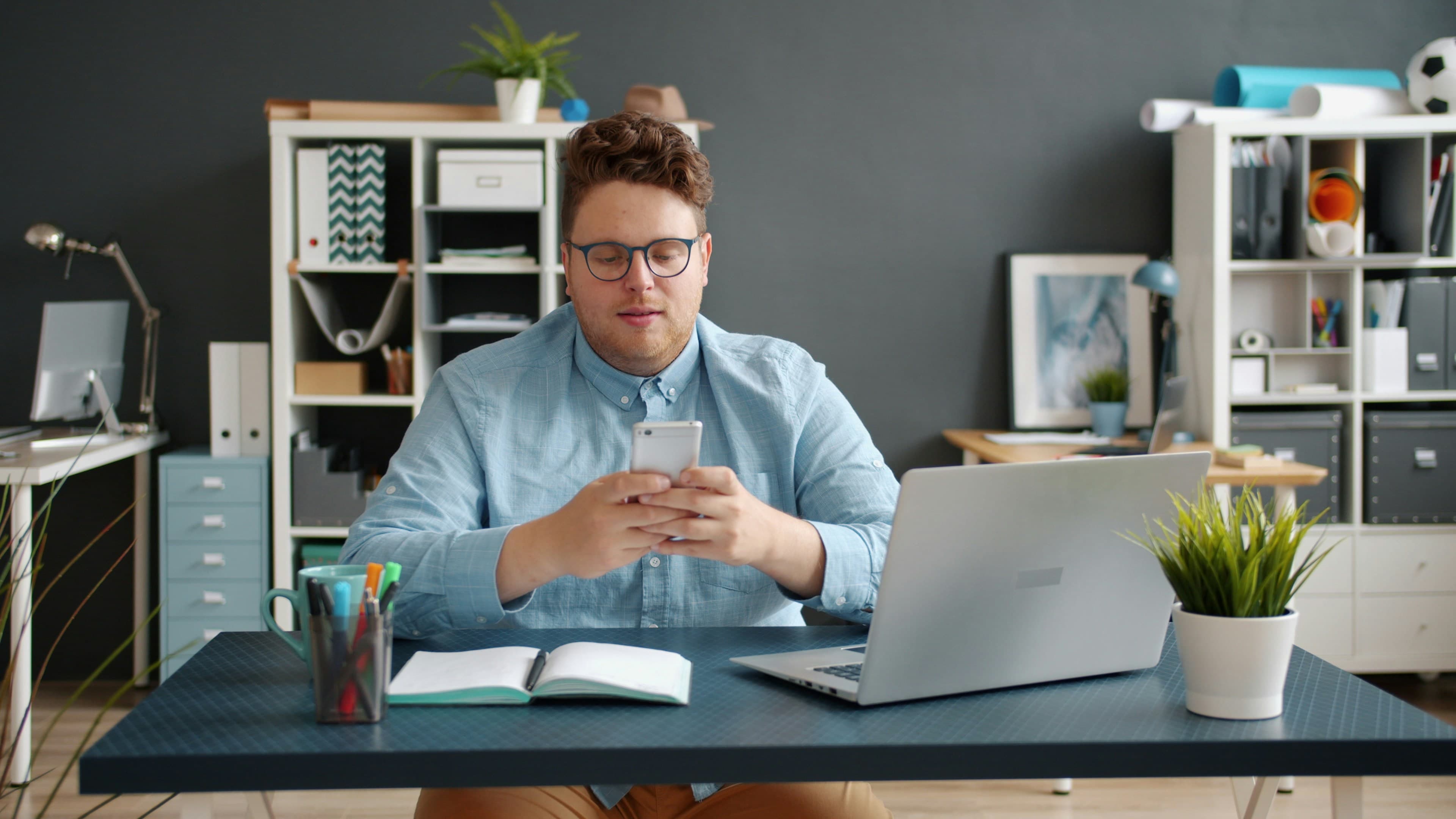 A man at his desk reading on his phone next to an open laptop